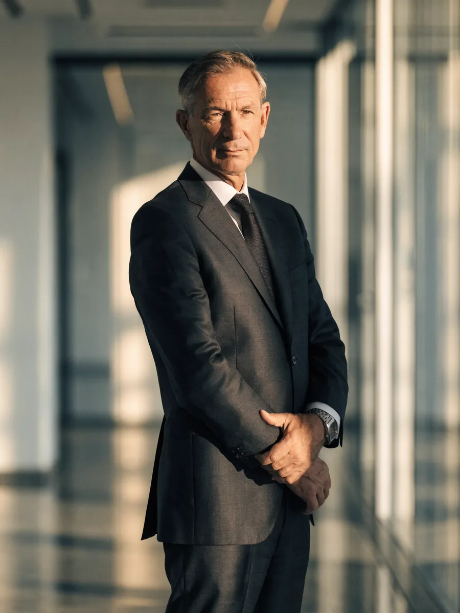 Senior business consultant in a charcoal suit standing with arms folded in a modern, naturally lit office corridor