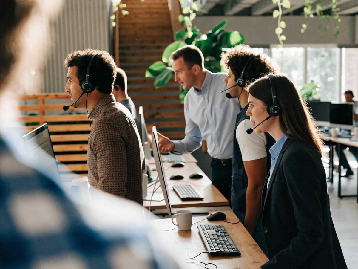 Sales team working at standing desks with headsets and monitors in a bright open-plan office while a team leader coaches a colleague