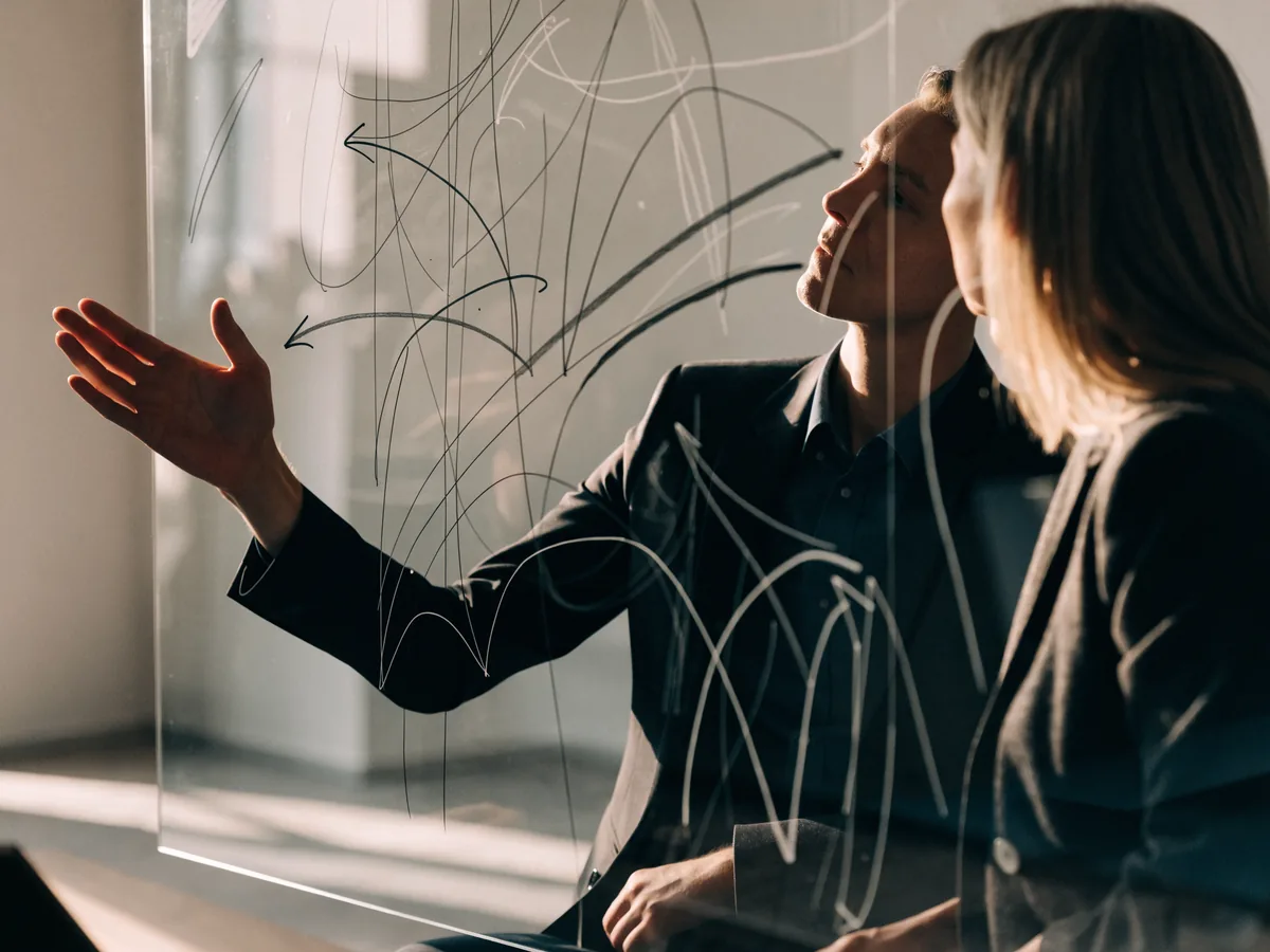 Two consultants mapping a growth strategy in diagrams and flow arrows across a large glass whiteboard in a minimal office